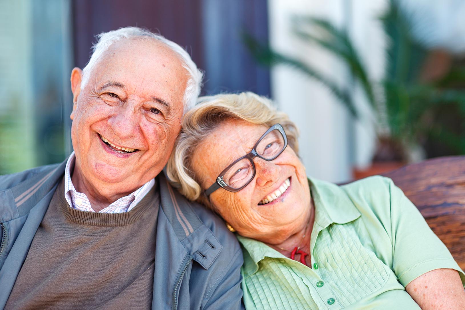 A senior couple is laughing. She has her head on his shoulder. She is wearing glasses and a green shirt. At her neck, she has a red pendant. He is wearing a blue jacket, a brown sweater and a white shirt. They are sitting on a wooden bench.